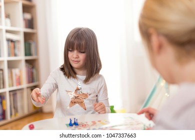 Mother And Daughter Sitting In A Playroom, Playing A Ludo Game; Daughter Rolling The Dice