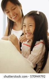 Mother And Daughter, Sitting Side By Side, Laughing While Mother Braids Daughter's Hair.