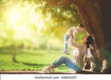 Mother And Daughter Sitting Under The Tree On Summer Lawn. Happy Family Playing Outdoors. Pretty Young Mom Having Fun With Her Baby In The Park Outside.