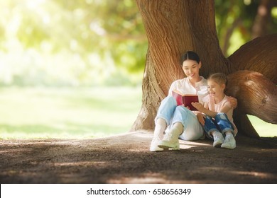 Mother And Daughter Sitting Under The Tree On Summer Lawn. Happy Family Playing Outdoors. Pretty Young Mom Reading A Book To Her Child In The Park Outside.