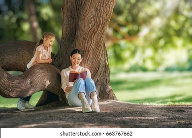 Mother And Daughter Sitting Under The Tree On Summer Lawn. Happy Family Playing Outdoors. Pretty Young Mom Reading A Book To Her Child In The Park Outside.