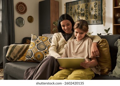 Mother and daughter smiling while using tablet together on a cozy living room sofa. Decorated in warm tones with patterned cushions and a tapestry wall hanging