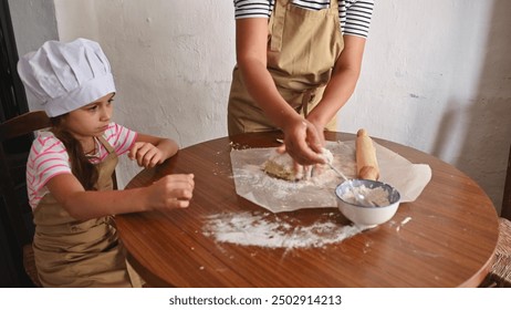 A mother and daughter spend quality time baking together. The child wears a chef hat and apron, watching intently as her mother prepares the dough on a wooden table. - Powered by Shutterstock