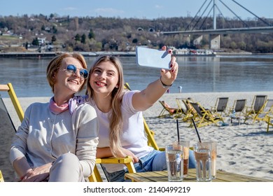 Mother and daughter spending quality time together, bonding, drinking coffee, and taking a selfie by the riverbank with the bridge in the background.