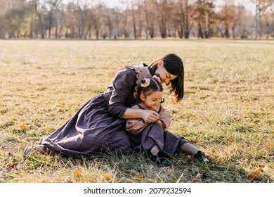 Mother And Daughter Spending Time Outdoors In A Park. Woman And Little Girl Sitting On Green Grass In A Field, Playing, Wearing Matching Outfits.