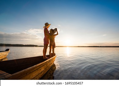 Mother And Daughter Stand Together Hugging On The Bow Of A Wooden Boat And Enjoy The Sunset By The Lake Or Sea. Summer Vacation Near The Water. Back View.