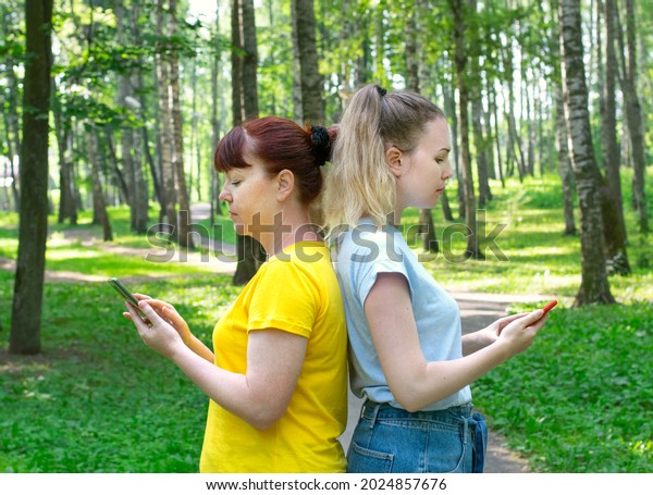 Mother and daughter are standing back to back\
using their phones in the park. Losing family connection because of\
social media and phone\
addiction