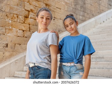 Mother And Daughter Standing On Stone Stairs Outside. A Middle-aged Woman And Kid Girl Holding Each Other Hands And Wearing Blue T-shirts And Looking Straight Into The Camera. Motherhood Concept