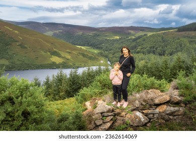 Mother and daughter standing and smiling on rock wall. Lake Lough Dan, forest and fields in background. Family hiking in Wicklow Mountains, Ireland
