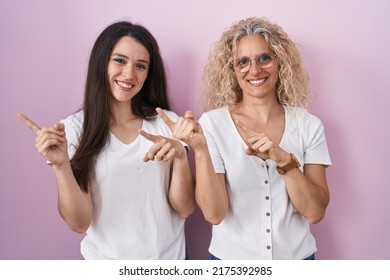 Mother And Daughter Standing Together Over Pink Background Smiling And Looking At The Camera Pointing With Two Hands And Fingers To The Side. 