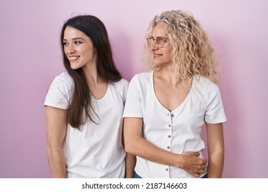 Mother And Daughter Standing Together Over Pink Background Looking Away To Side With Smile On Face, Natural Expression. Laughing Confident. 