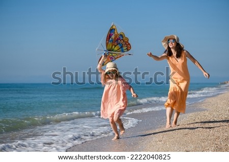 Mother and daughter in summer dresses and hats fly kite at sea beach coast , active happy family vacation
