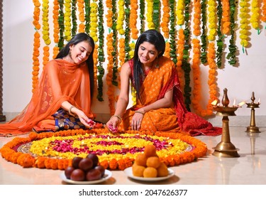 Mother and daughter in tradional clothes making rangoli at home on the occasion of deepawali festival with diya lamps ,flowers and mithai