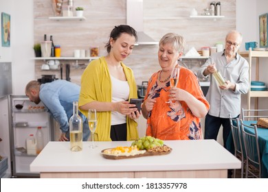 Mother and daughter using smarthphone in kitchen. Senior woman holding glass of wine. Old man looking at bottle. - Powered by Shutterstock