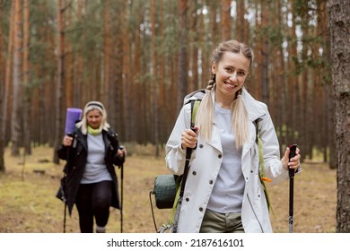 Mother And Daughter Walking In The Forest. Young Girl Is Walking Ahead, Elderly Woman Behind. Women Are Holding Rucksack, Trecking Poles. Woman Is Smiling.