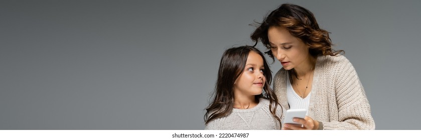 Mother And Daughter In Warm Knitwear Looking At Each Other Near Smartphone Isolated On Grey, Banner