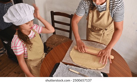 Mother and daughter wearing aprons and chef hats, rolling dough on a table in a cozy kitchen. Family bonding and baking at home. - Powered by Shutterstock