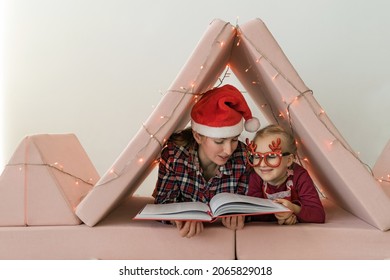 Mother with daughter wearing pajamas and Christmas hat and fun glasses reading book in baby room. Closeup of woman and little girl relaxing in DIY play tent decorated with festive light on Christmas - Powered by Shutterstock