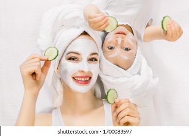 Mother And Daughter In White Shirts And White Towels On Their Heads In A Home Bathroom, Doing Spa Procedures With Mud Mask And Fresh Cucumber Slices. Family Time, Spa And Beauty, Mothers Day