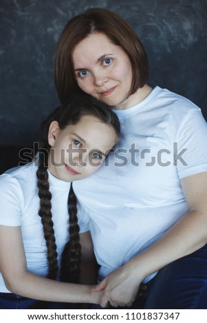 Mother and daughter in white T-shirts and blue jeans, hugging and smiling in the studio on a dark gray brick background.The concept of style casual.