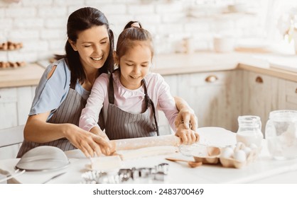 A mother and daughter work together in a kitchen, rolling out dough for cookies. The mother has her arm around the daughter and is guiding her hand as she uses a rolling pin - Powered by Shutterstock