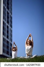 Mother and daughter working out practicing yoga outside on a grass together urban city  park on blue sky background