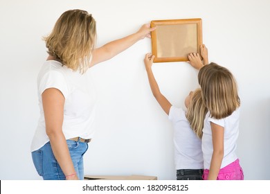 Mother And Daughters Hanging Blank Photo Frame On White Wall. Back View Of Blonde Mom Holding And Aligning Empty Picture With Help Of Two Kids. Family, Relocation And Moving Day Concept