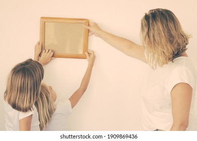 Mother And Daughters Hanging Blank Photo Frame On White Wall. Back View Of Blonde Mom Holding And Aligning Empty Picture With Help Of Two Kids. Family, Relocation And Moving Day Concept