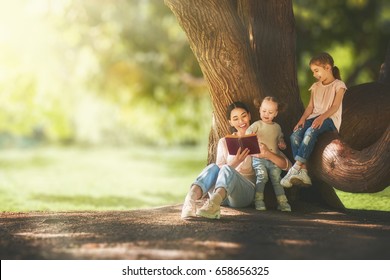 Mother And Daughters Sitting Under The Tree On Summer Lawn. Happy Family Playing Outdoors. Pretty Young Mom Reading A Book To Her Children In The Park Outside.