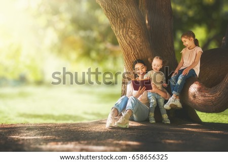 Mother and daughters sitting under the tree on summer lawn. Happy family playing outdoors. Pretty young mom reading a book to her children in the park outside.