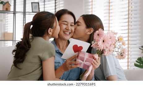 Mother day two grown up child cuddle hug give flower gift box red heart card to mature mum. Love kiss care mom asia middle age adult three people sitting at home sofa happy smile enjoy family time. - Shutterstock ID 2279311691