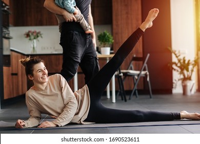 Mother Doing Side Leg Raises While Father Walks Around, Carrying Their Infant Baby In His Hands. She Looks At Camera, Laughing, Amused.Family Quarantine, Domestic Life In Self-isolation. Sunset Light