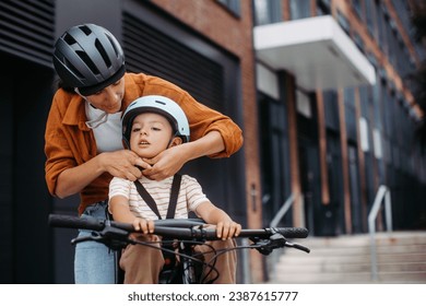 Mother fastening sons' bike helmet on head, carring him on child bike carrier, seat. Mom commuting with a young child through the city on a bicycle. - Powered by Shutterstock