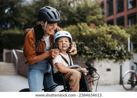 Mother fastening sons' bike helmet on head, carring him on child bike carrier, seat. Mom commuting with a young child through the city on a bicycle.