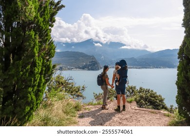 Mother and father with baby carrier looking at lakeside town Nago Torbole seen from hiking trail Sentiero del Ponale at Garda Lake, Trentino, North Italy. Surrounded by mountains of Garda Hills - Powered by Shutterstock