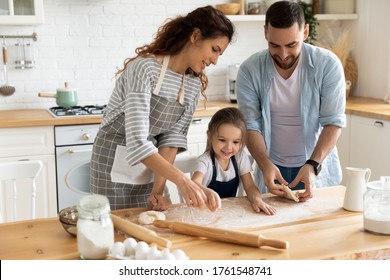 Mother And Father With Cute Little Daughter Cooking Organic Flour Standing At Table In Kitchen. Smiling Caring Mum And Dad With Girl Using Rolling Pin For Pastry On Wooden Board, Preparing Dinner.