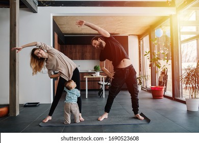 Mother And Father Doing Side Tilts As A Part Of Their Morning Exercise, While Their Child Pulling Mother's Shirt, Seeking Attention. Family Quarantine, Domestic Life In Self-isolation. Sunset Light