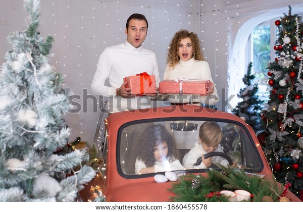 Mother and\
father with gift boxes in their hands and near are theis children\
in red car near Christmas\
trees.