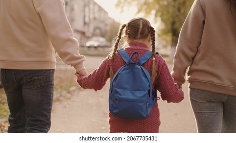 Mother And Father Hold A Little Schoolgirl With A Backpack By The Hand, Happy Family, Walk Through Schoolyard In Fall, Mom, Dad And Kid With Bag Over Their Shoulders, Accompany Child To First Grade