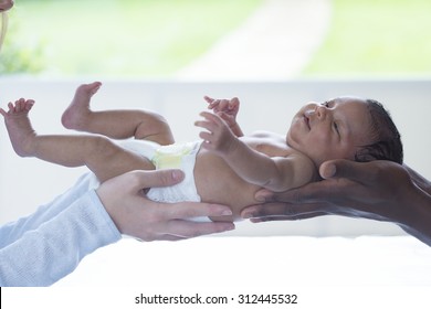 A Mother And Father Holding A Newborn Baby. The Baby Is Wearing A Nappy And Only The Mother's And Father's Hands Can Be Seen. 