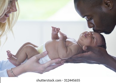 Mother And Father Holding Their New Born Baby. The Mother Is Smiling And The Father Is Kissing The Baby's Head.