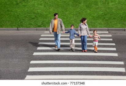 mother and father holds hand of little daughter and son and crossing road, green fence and grass