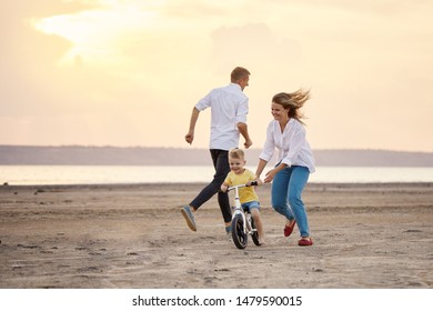 Mother And Father With Kids Riding Scooter And Bike In Summer. Family Runs On The Beach At Sunset. Daughter And Son Learns To Ride