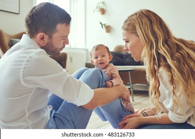 Mother And Father Looking Concerned At Their Crying Newborn Baby Daughter, Sitting On The Floor At Home