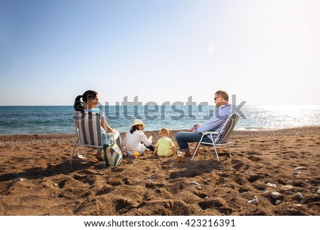 Mother and father are sitting on a beach deck chair, daughter and son playing
