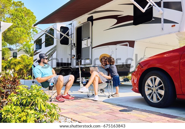Mother,\
father and son sitting near camping trailer,smiling.Woman, men, kid\
relaxing on chairs near car and palms.Family spending time together\
on vacation near sea or ocean in modern rv\
park