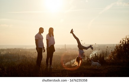 Mother and father are standing on the hill and watching their daughter doing a handstand, behind them beautiful view horizon of a setting sun - Shutterstock ID 1676235358