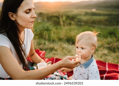 Mother feed baby boy with yogurt and sits on blanket on a picnic in mountains. Mom play with son in sunset. Happy family holiday on summer day. Parents with child. Spend time together. - Powered by Shutterstock