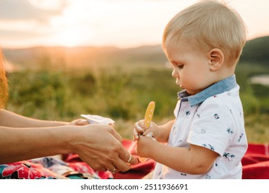 Mother feed baby boy with yogurt and sits on blanket on a picnic in mountains. Face closeup. Mom play with son in sunset. Happy family holiday on summer day. Parents with child. Spend time together. - Powered by Shutterstock