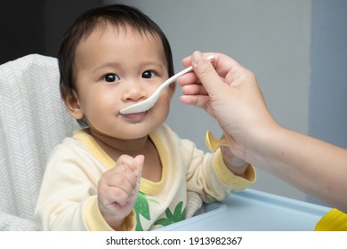 
Mother Feed Little Toddler Baby Boy With Open Mouth From Plastic Spoon In High Chair View From Back